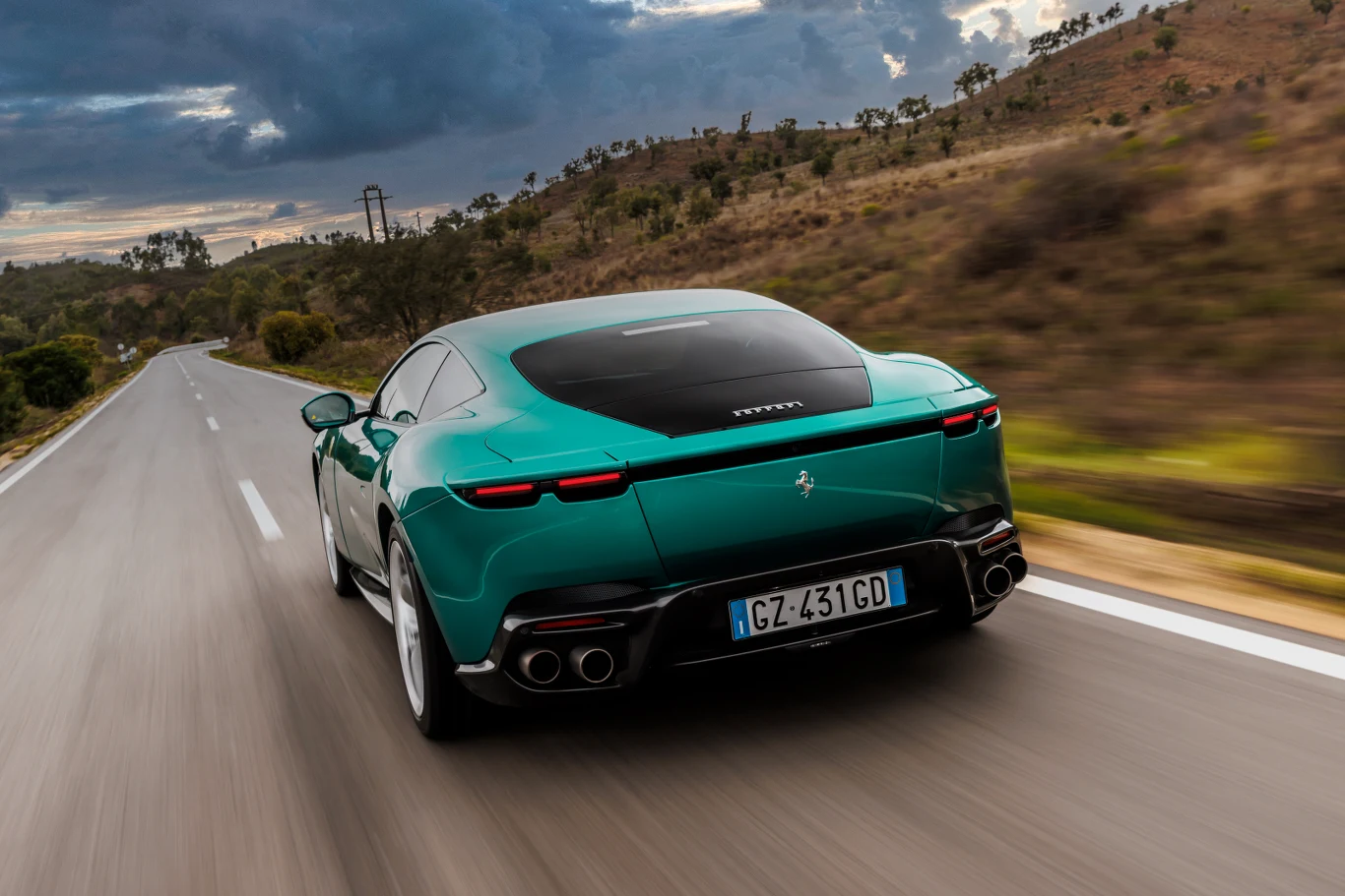 Zielony sportowy samochód marki Ferrari jadący pustą, wąską drogą pośród pagórkowatego krajobrazu, widok od tyłu na pojazd poruszający się dynamicznie w kierunku horyzontu, po lewej stronie zadrzewione wzgórza.