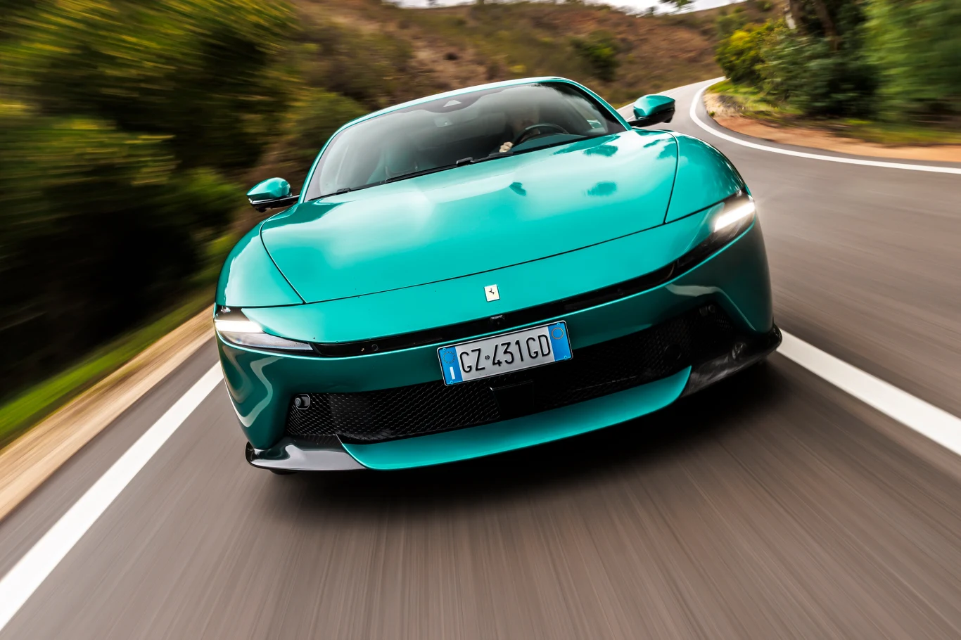 Jasnozielone Ferrari poruszające się dynamicznie po krętej drodze asfaltowej otoczonej zielenią, uchwycone z perspektywy z przodu, podkreślającej kształty i sportowy charakter samochodu.