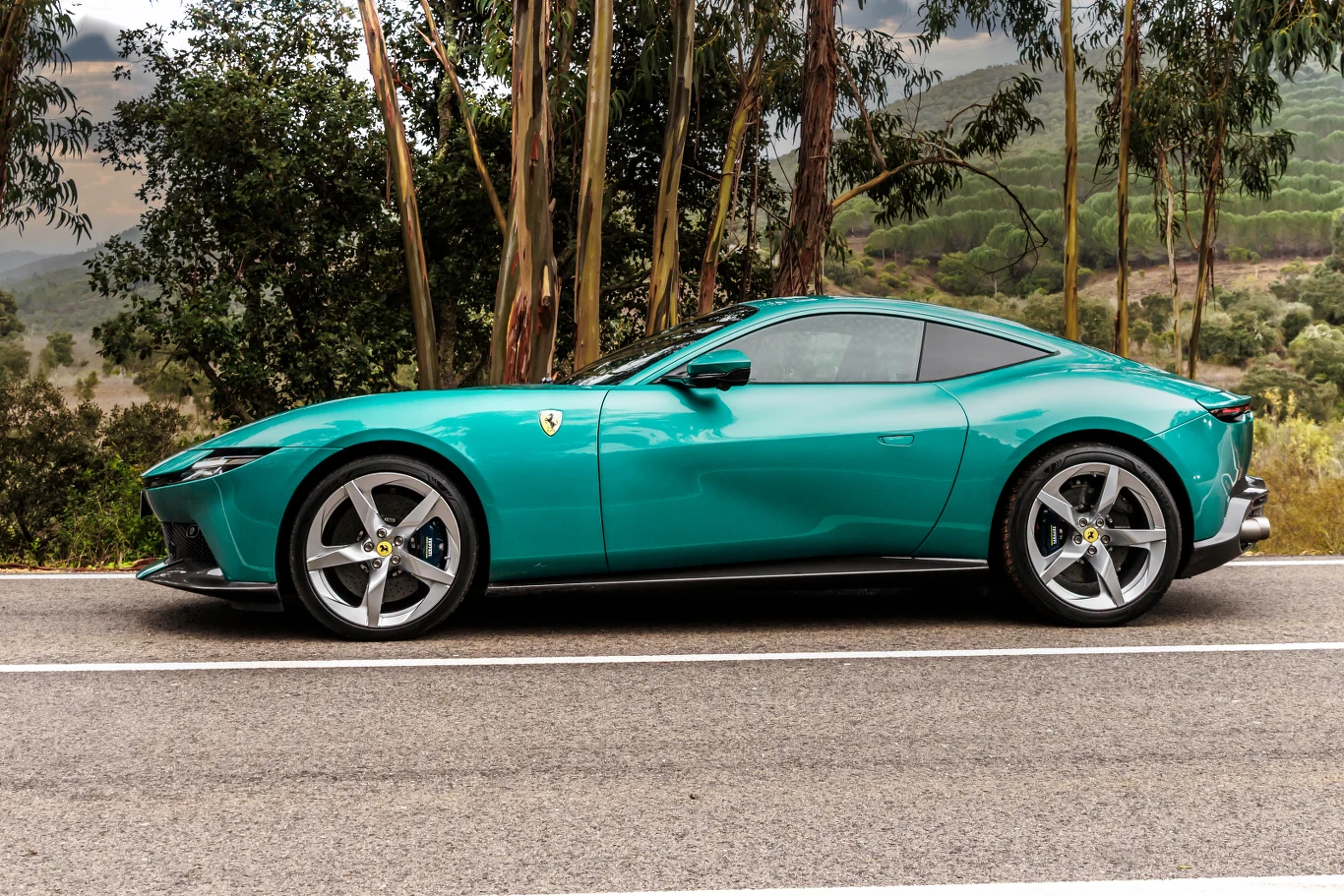 Nowoczesny sportowy samochód Ferrari w kolorze turkusowym zaparkowany przy drodze, wokół roślinność śródziemnomorska, w tle wzgórza i drzewa.