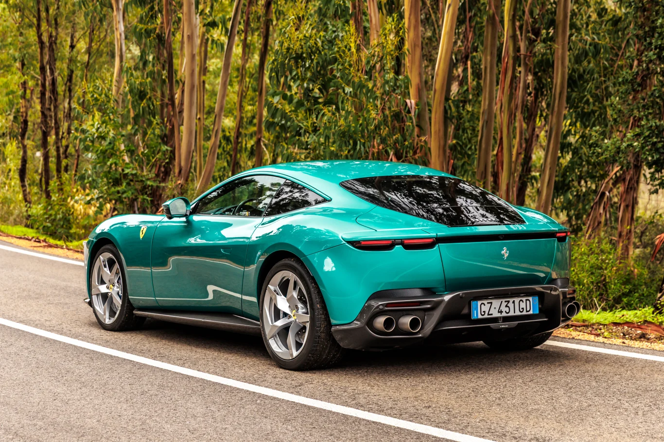 Sportowy samochód Ferrari w zielonym kolorze parkuje na poboczu asfaltowej drogi otoczonej wysokimi drzewami. Tylne światła oraz charakterystyczne, podwójne rury wydechowe dodają nowoczesnego, dynamicznego wyglądu.