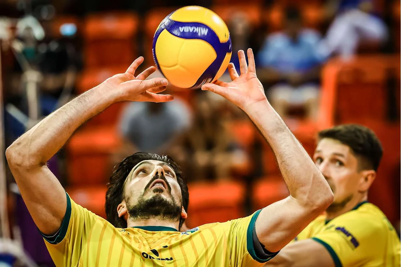 Zawodnik w żółtej koszulce siatkarskiej unosi ręce, przygotowując się do precyzyjnego wystawienia piłki podczas rozgrywki. W tle widać drugiego zawodnika i rozmyte trybuny.