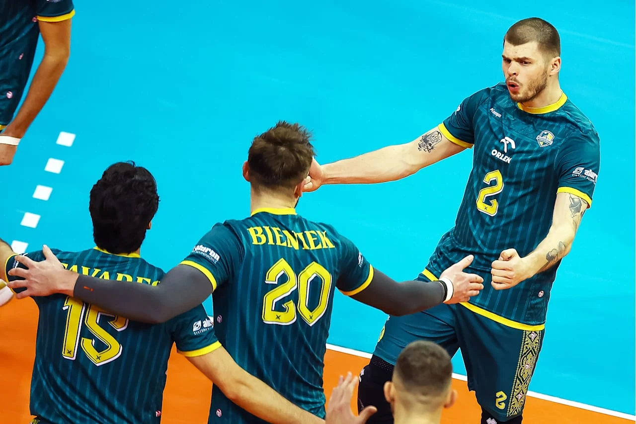 Siatkarze w zielonych strojach drużynowych świętują zdobycie punktu na hali sportowej, przy czym na pierwszym planie widać dynamiczne gesty radości i napięcia emocjonalnego.