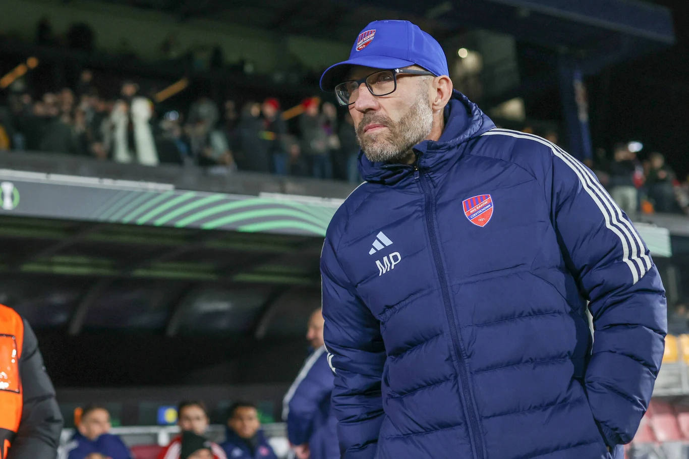 Mężczyzna w niebieskiej kurtce sportowej i czapce z daszkiem z herbem klubu piłkarskiego, otoczony przez innych ludzi na trybunach stadionu podczas wydarzenia sportowego.