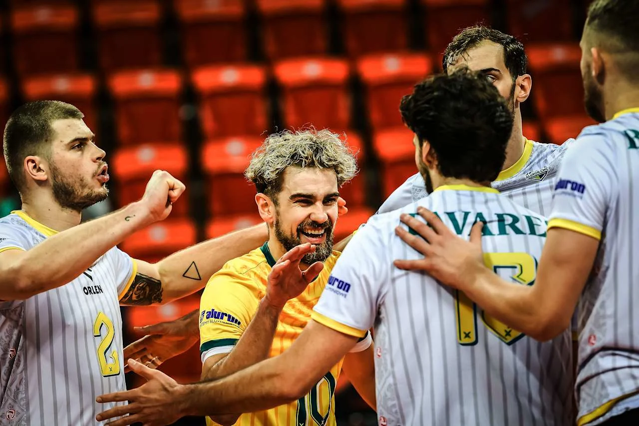 Siatkarze Aluron CMC Warty Zawiercie zagrają teraz z zespołem z Japonii Grupa siatkarzy w sportowych strojach świętuje wspólny sukces na boisku. Centralnie uśmiechnięty zawodnik w żółtej koszulce otoczony przez kolegów z drużyny wyrażających radość. W tle czerwone trybuny.