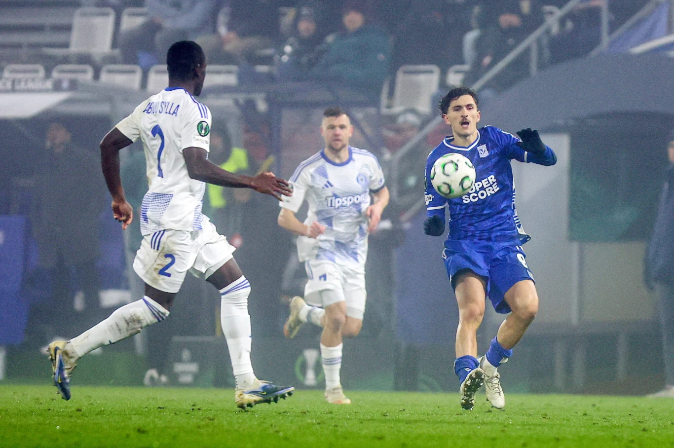 Abdoulaye Sylla i Pablo Rodriguez (z prawej) w meczu Sigma - Lech Trzech piłkarzy biegnie po boisku podczas rozgrywki piłkarskiej, zawodnik w niebieskiej koszulce kontroluje piłkę, dwóch zawodników drużyny przeciwnej w białych strojach obserwuje sytuację. W tle widoczne są trybuny i kibice.