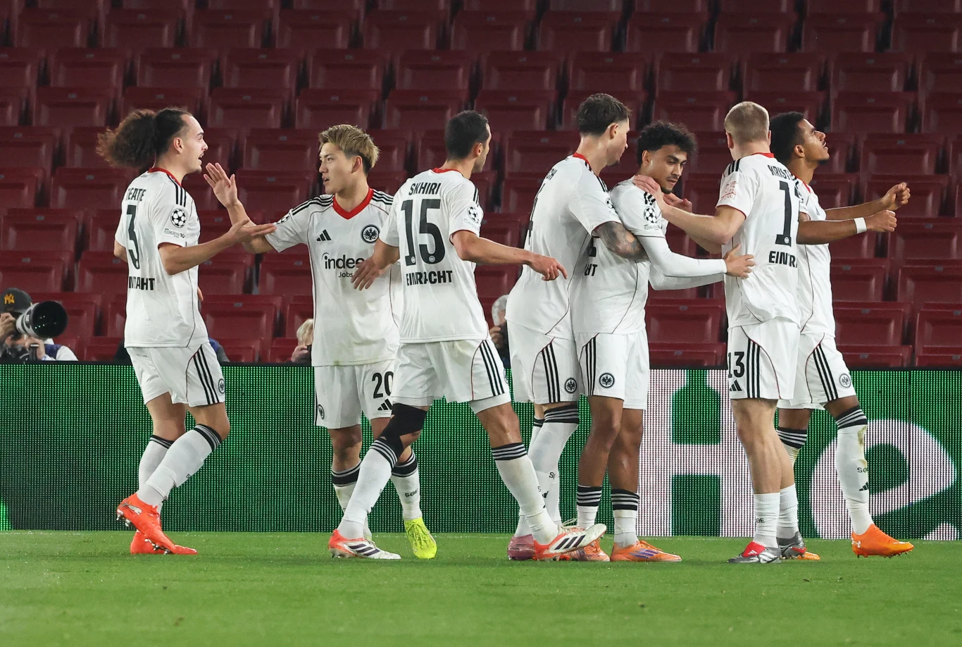 Grupa piłkarzy w białych strojach świętuje zdobycie gola, stojąc na murawie stadionu. W tle widoczne są puste czerwone trybuny oraz fragmenty reklam cyfrowych przy linii bocznej.