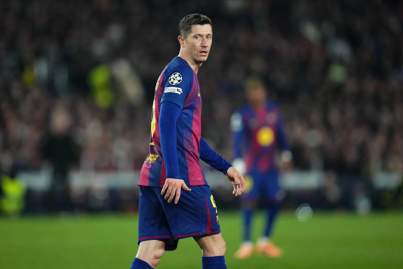 Piłkarz ubrany w granatowo-bordowy strój ligowy spaceruje po murawie stadionu w trakcie meczu. W tle widoczni są inni zawodnicy oraz rozmazana publiczność na trybunach. Piłkarz nosi opaskę na ramię.