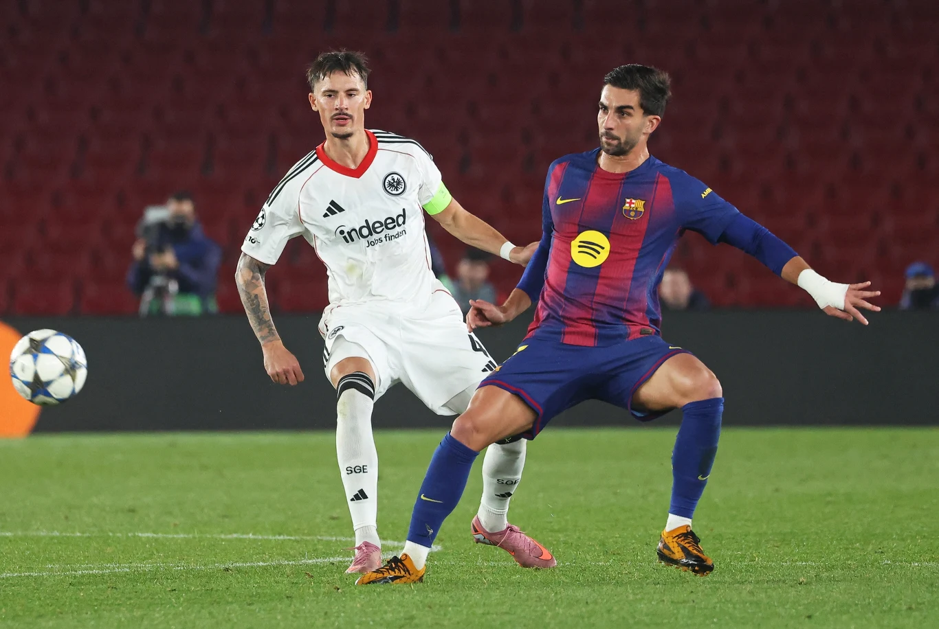 Dwóch zawodników rywalizuje o piłkę na boisku, jeden z nich w białym stroju z logo Eintracht Frankfurt, drugi w granatowo-bordowym stroju Barcelony. Trybuny w tle są słabo zapełnione, a uwagę zwracają dynamiczne pozy piłkarzy.