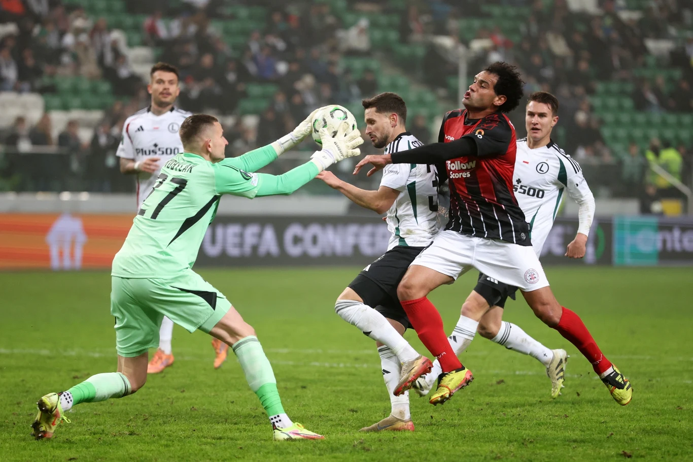 Bramkarz w jasnozielonym stroju wyciąga ręce po piłkę w trakcie dynamicznej akcji na boisku piłkarskim; wokół niego walczy kilku zawodników obu drużyn, w tle rozmazane trybuny stadionu.