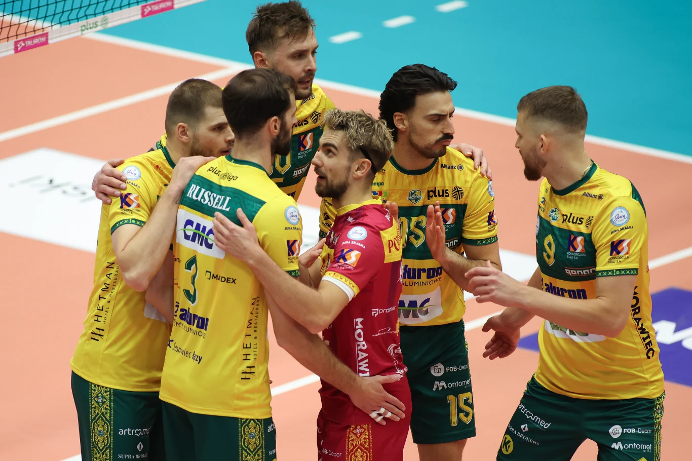 Grupa siatkarzy w żółto-zielonych strojach zbiera się w kręgu podczas przerwy w meczu, negocjując strategię i wzajemnie się motywując. Na środku widoczny jest libero w czerwonym stroju.