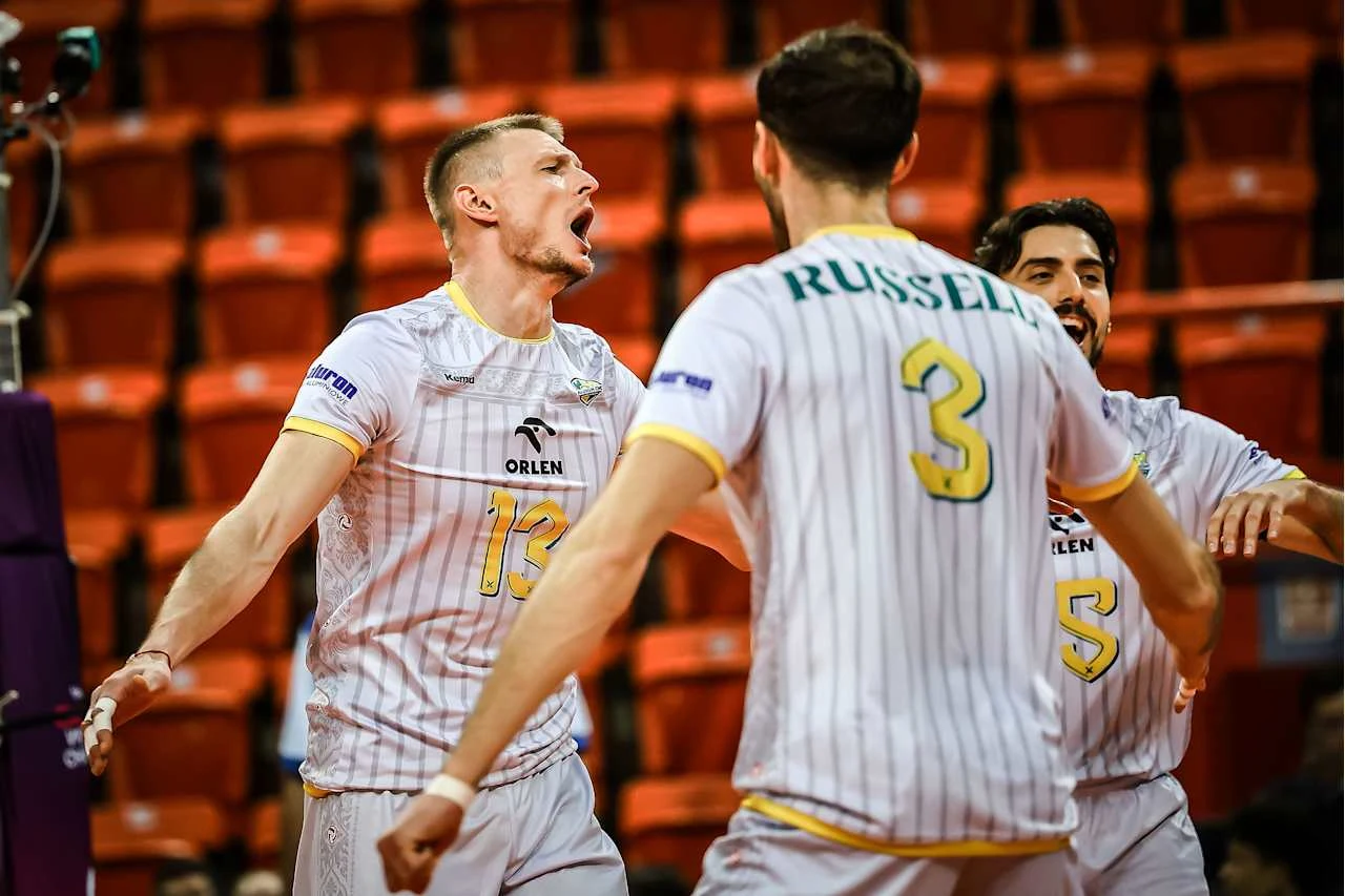 Siatkarze Aluron CMC Warty Zawiercie są w Brazylii niepokonani Grupa siatkarzy w białych strojach z żółtymi numerami na tle pustych, pomarańczowych trybun. Zawodnik oznaczony numerem 13 wyraża emocje, szeroko otwierając usta, podczas gdy zawodnik z numerem 3 stoi do niego tyłem. Trzeci zawodnik jest lekko uśmiechn...