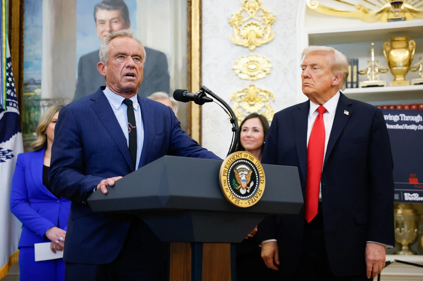 Administracja Trumpa zamierza wstrzymać płatności za zabiegi korekty płci u młodych Amerykanów Mężczyzna w garniturze przemawia do mikrofonu przy mównicy z pieczęcią prezydenta Stanów Zjednoczonych, obok niego stoi inny mężczyzna w garniturze oraz dwie kobiety w tle. W pomieszczeniu widoczne są złote dekoracje i portret na ścianie.