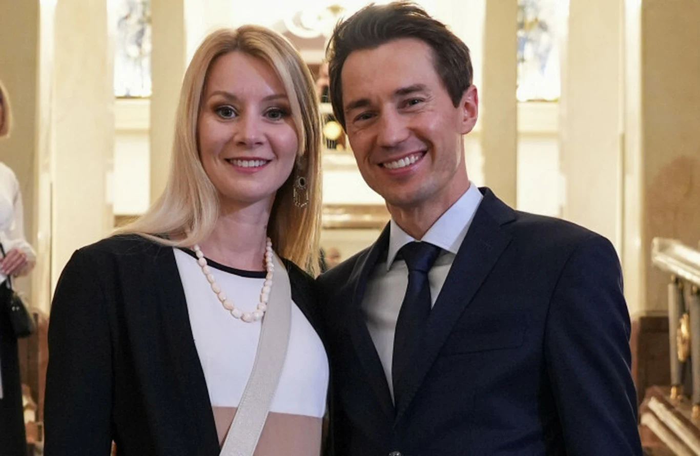 Ewa Bilan-Stoch, Kamil Stoch Para elegancko ubranych osób stojąca razem w jasnym pomieszczeniu z rozświetlonym tłem, kobieta ma na sobie czarną marynarkę i biały naszyjnik, mężczyzna w garniturze z krawatem, oboje uśmiechnięci, atmosfera formalna.