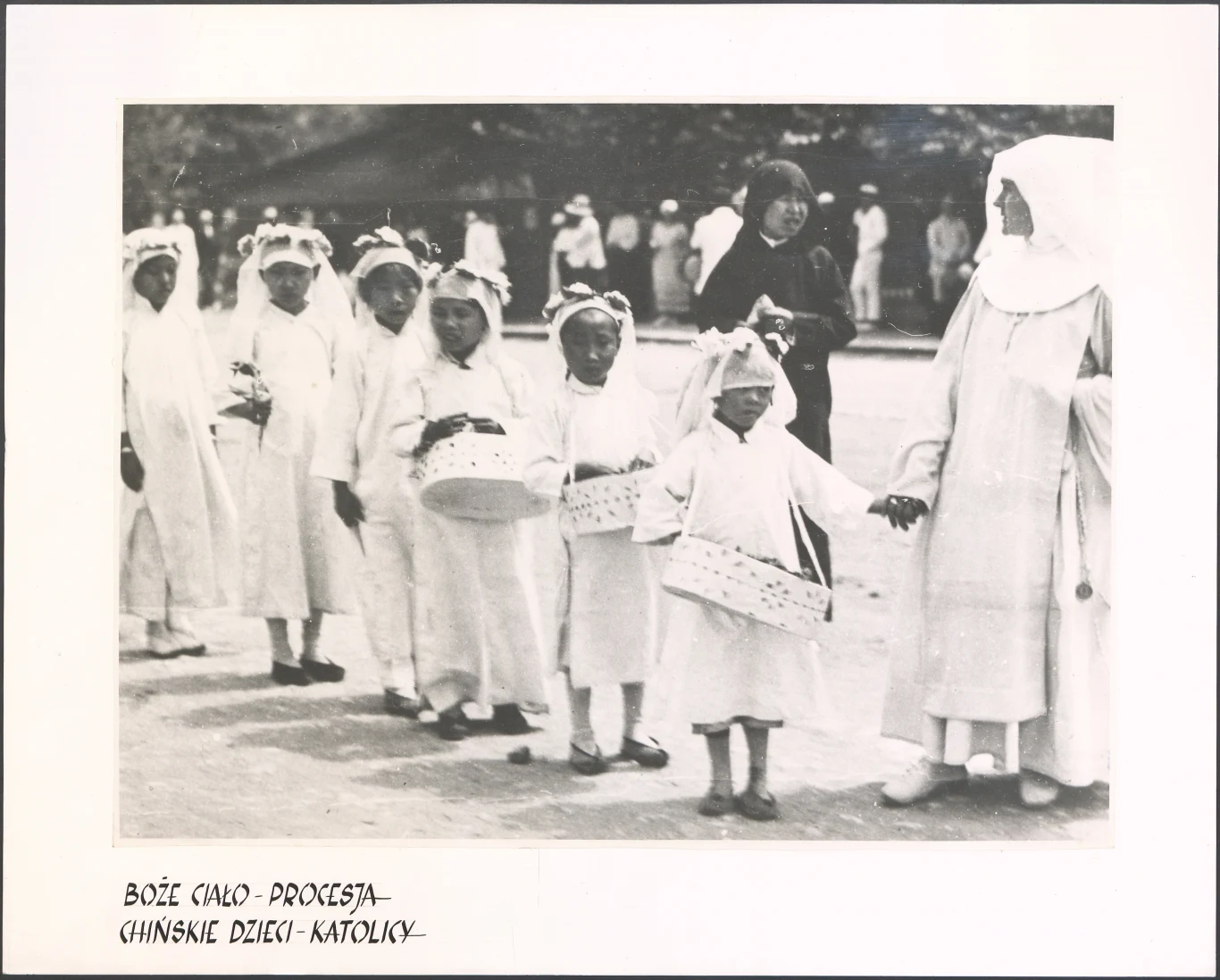 Grupa chińskich dzieci ubranych w białe uroczyste stroje, trzymających koszyczki, stoi w szeregu podczas procesji, obok nich zakonnica w białym habicie.