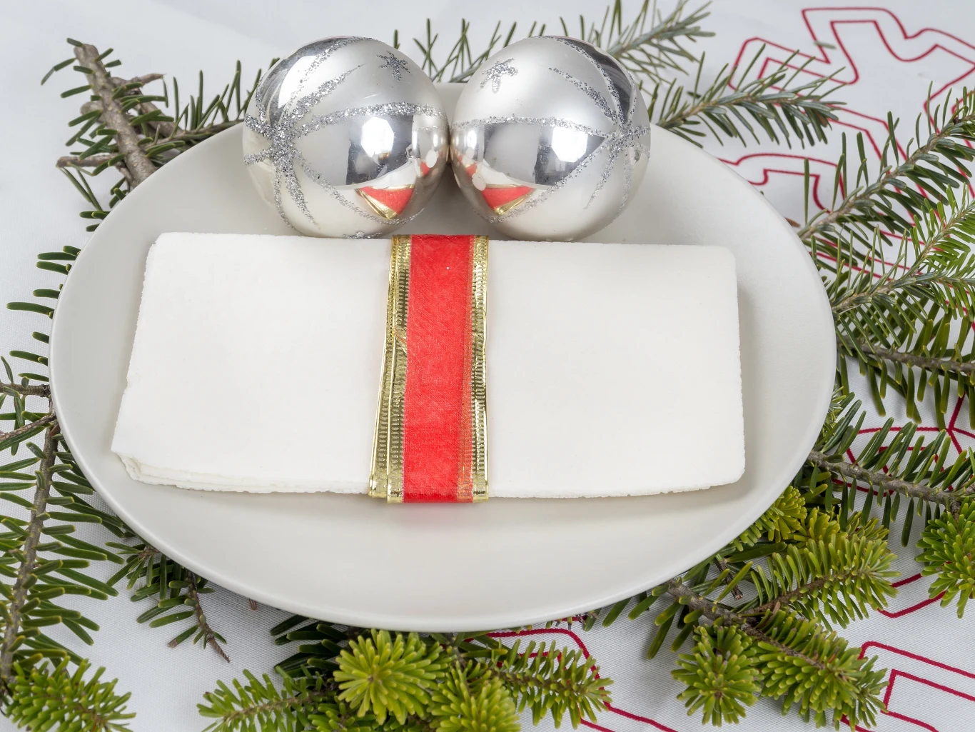 Porcja białego opłatka z czerwoną i złotą wstążką na talerzu, srebrne bombki oraz gałązki świerku na białym obrusie z haftem, dekoracja świąteczna.