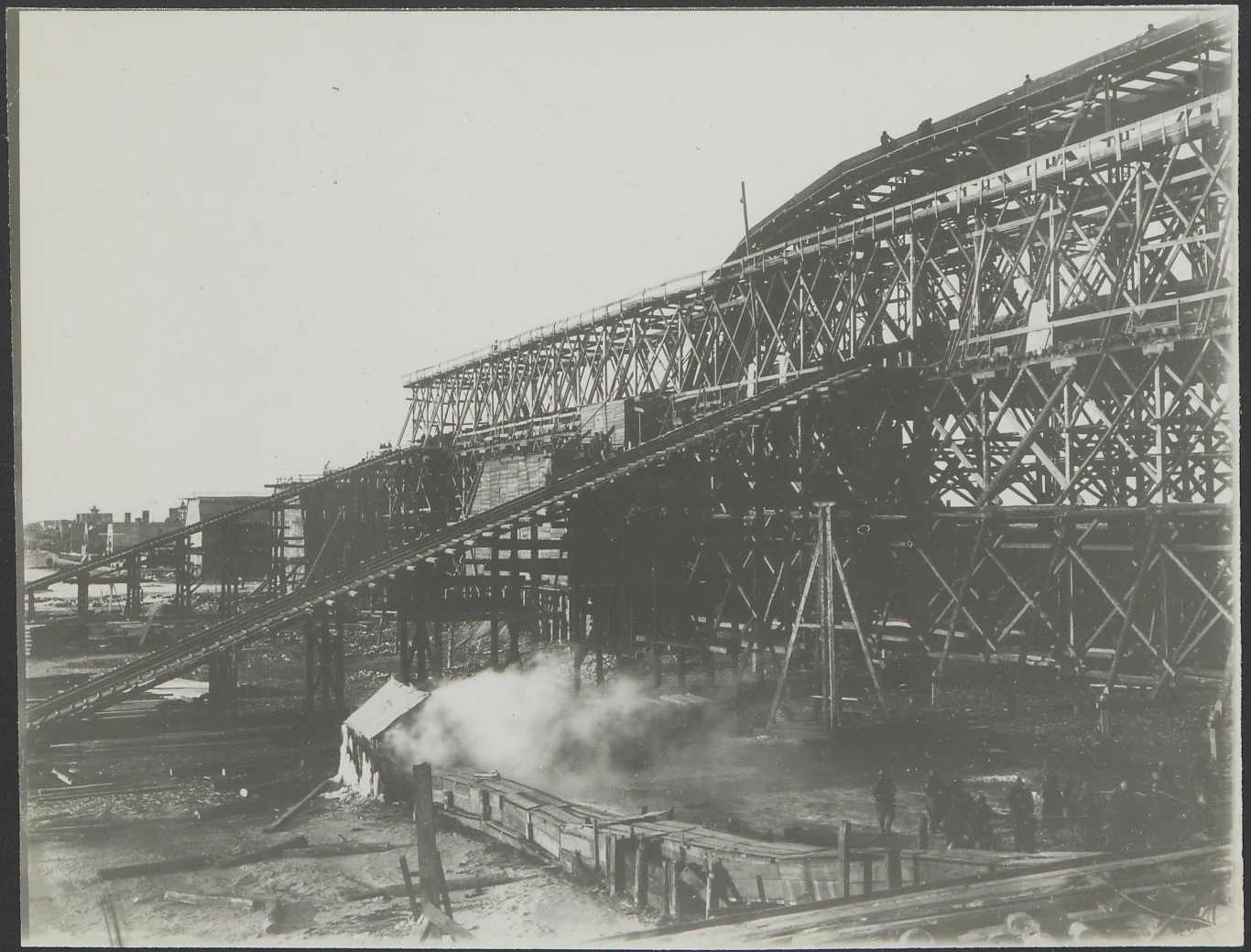 Wielka konstrukcja stalowa na placu budowy, składająca się z licznych rusztowań i podestów. Na pierwszym planie widać mniejszą zabudowę oraz dym lub parę unoszącą się z niewielkiego obiektu. Pracownicy są widoczni w tle blisko rusztowań.