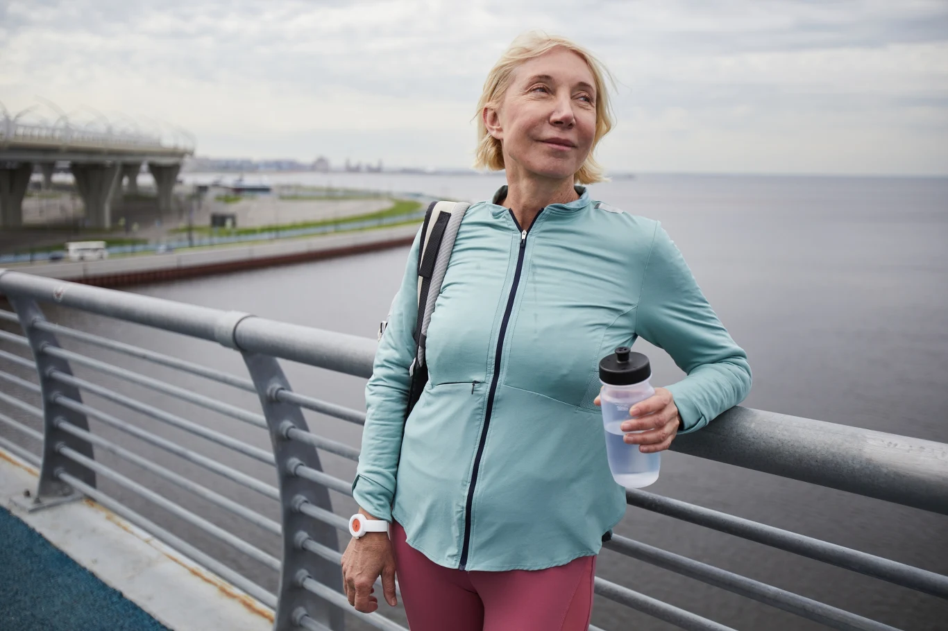 Zdrowe serce zaczyna się od codziennego ruchu, dostępnego dla większości z nas Starsza kobieta w sportowym stroju opiera się o barierkę nad wodą, w dłoni trzyma butelkę na wodę, na jej twarzy widoczny jest spokojny wyraz, a w tle znajduje się nowoczesna infrastruktura mostu.