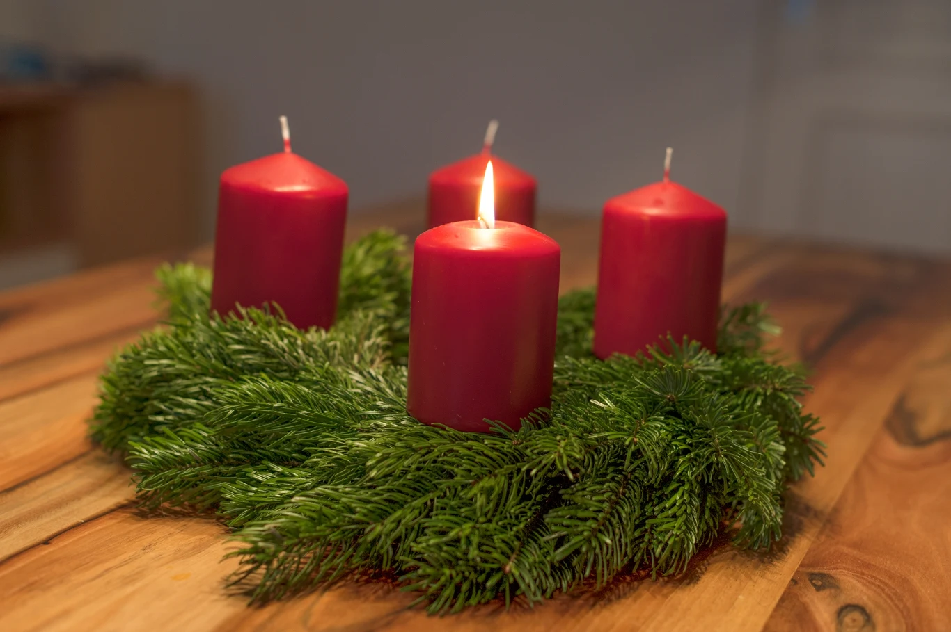 Cztery czerwone świece ustawione na zielonym wieńcu z gałązek iglastych, z których jedna świeca jest zapalona, całość leży na drewnianym stole.