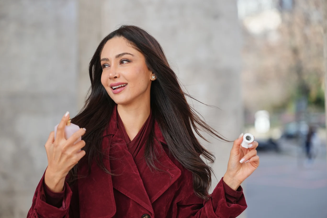 Kobieta o długich ciemnych włosach ubrana w bordowy płaszcz, uśmiechnięta, trzymająca w dłoniach flakonik perfum oraz zakrętkę, rozpyla zapach na tle jasnej ściany.