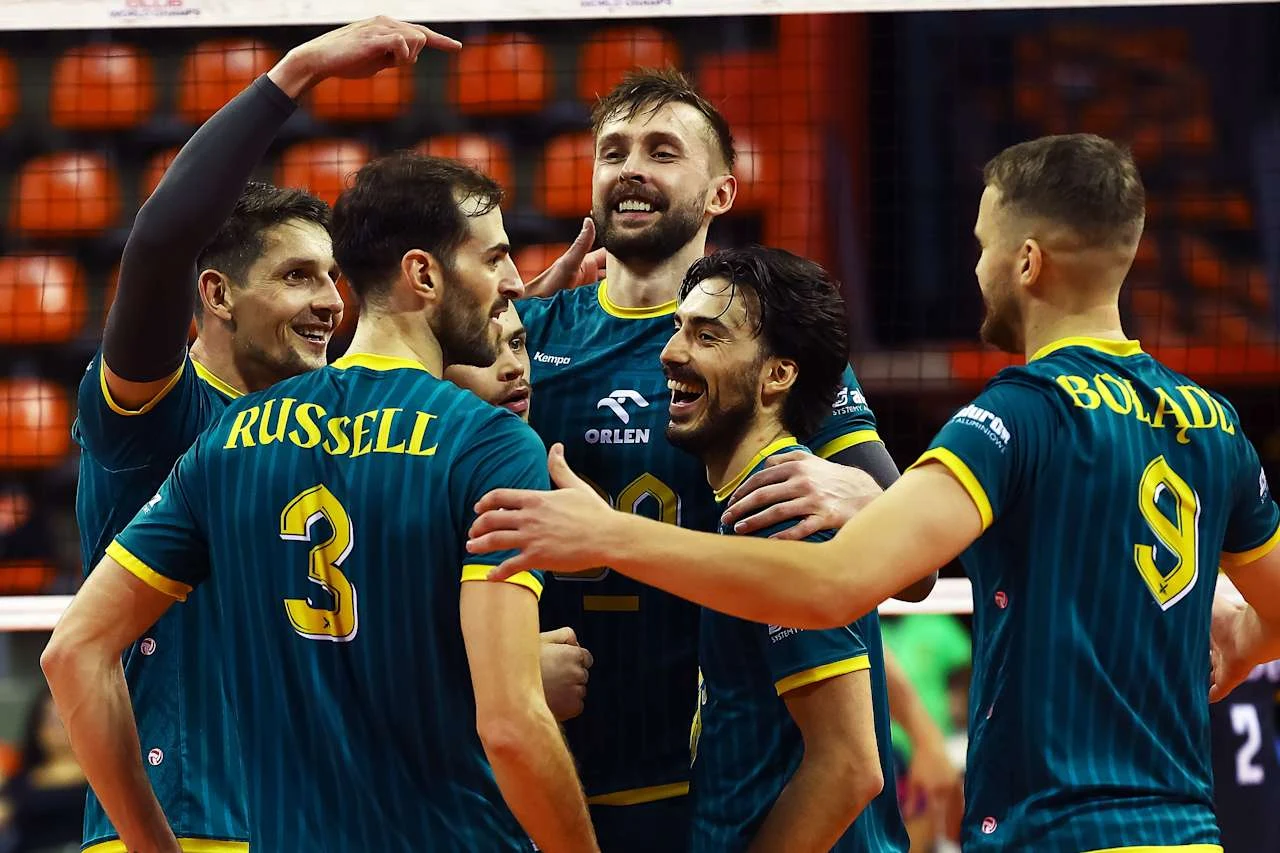 Siatkarze Aluron CMC Warty Zawiercie Grupa siatkarzy w zielonych strojach z żółtymi akcentami cieszy się wspólnie po zdobyciu punktu na boisku sportowym, w tle widać trybuny i siatkę.