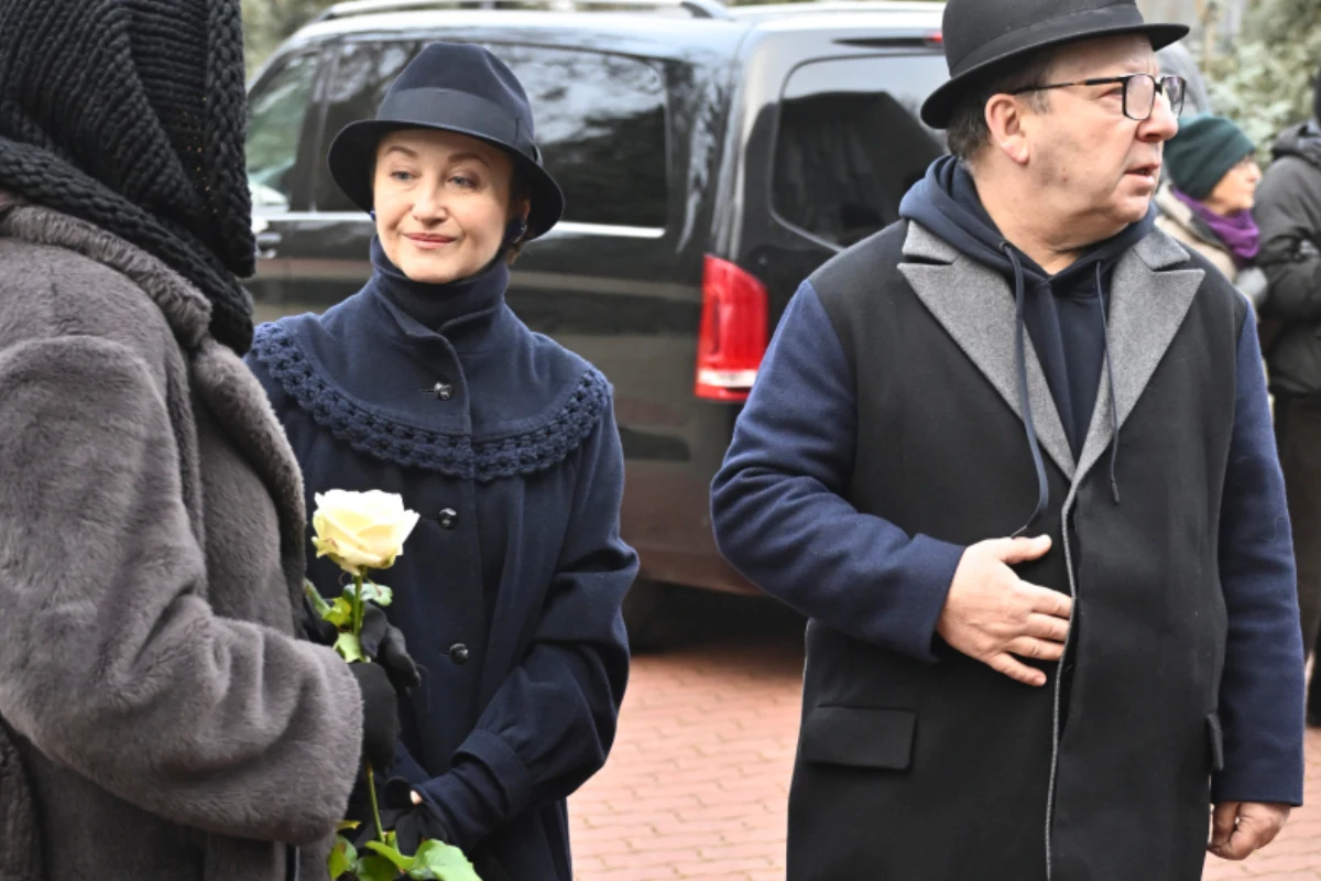 Grupa dorosłych ubranych w ciemne, eleganckie stroje, prawdopodobnie uczestnicząca w uroczystości pogrzebowej. Jedna z osób trzyma białą różę, a w tle stoi czarny samochód.