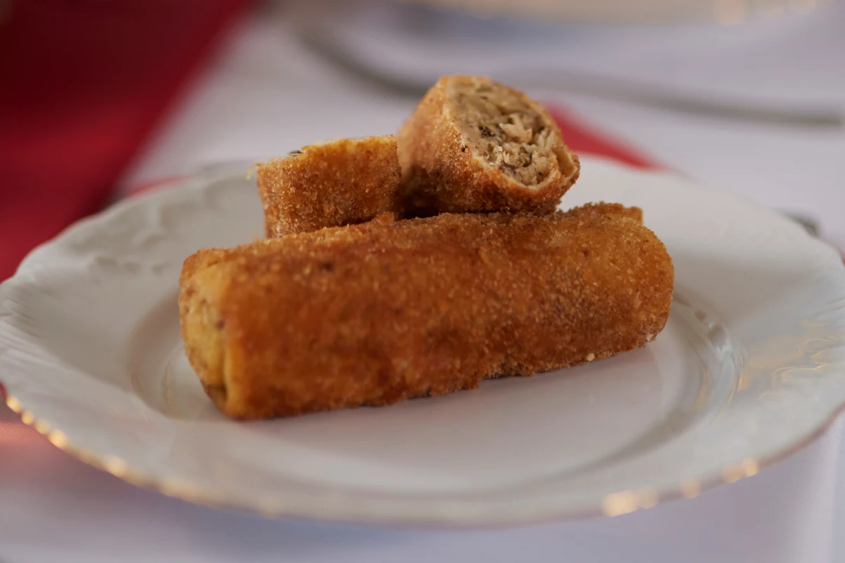 Dwa złociste krokiety z farszem mięsnym na białym, porcelanowym talerzu, jeden przekrojony ukazujący wnętrze, w tle rozmyta tkanina koloru czerwonego.