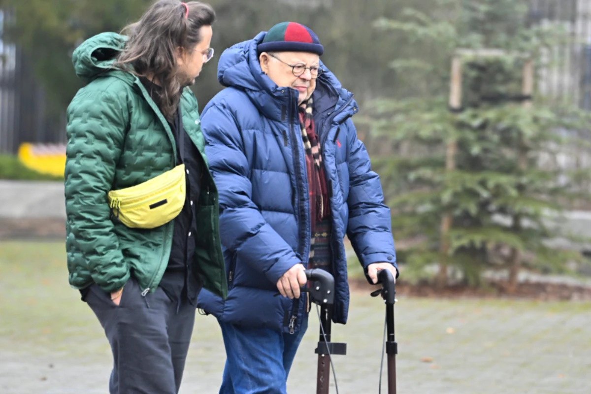 Dwóch mężczyzn spacerujących alejką w parku, starszy pan wspiera się na dwóch laskach, obok idzie młodszy mężczyzna w zielonej kurtce z żółtą saszetką, panuje chłodna pogoda, obaj mają na sobie ciepłe ubrania.