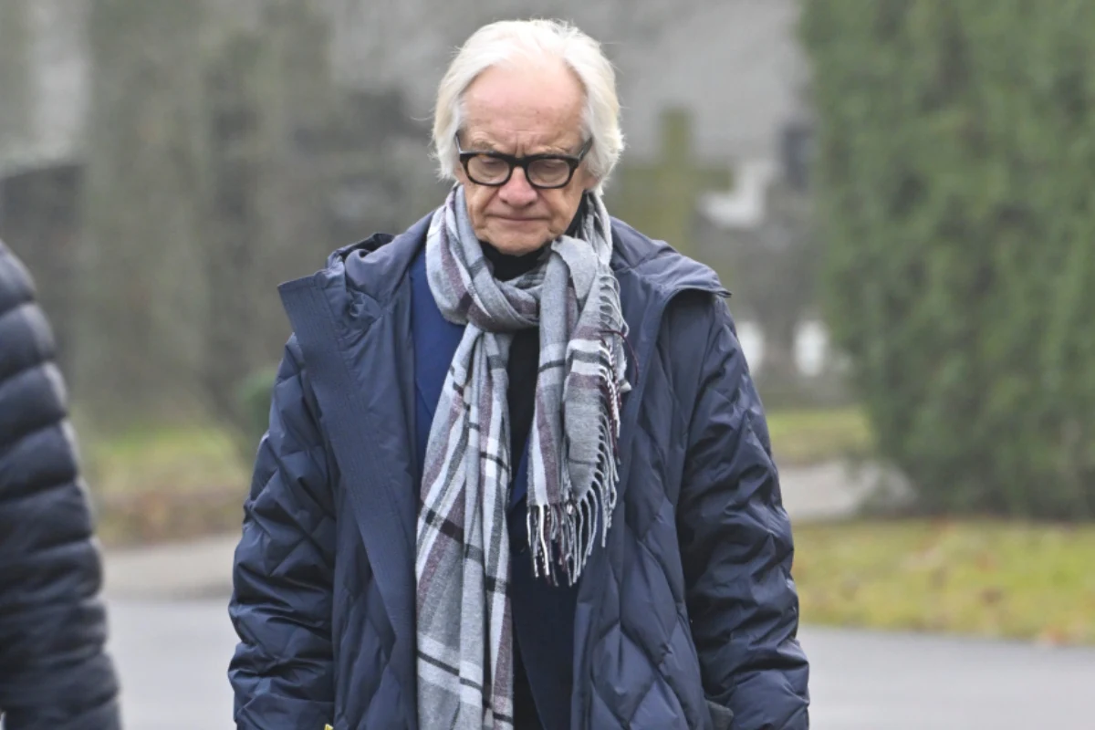 Starszy mężczyzna z siwymi włosami i okularami ubrany w ciemną kurtkę oraz szalik, stoi na zewnątrz w pochmurny dzień. W tle widoczne rozmyte drzewa i fragmenty nagrobków, sugerujące cmentarz.