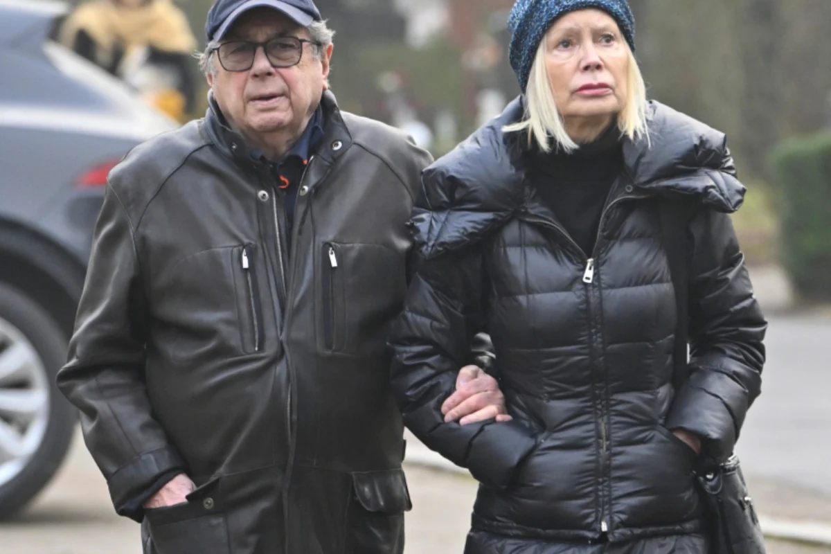 Starszy mężczyzna i kobieta spacerują razem, ubrani w zimowe kurtki. Kobieta ma blond włosy i nosi niebieską czapkę, mężczyzna ma okulary, czapkę z daszkiem i ręce w kieszeniach.