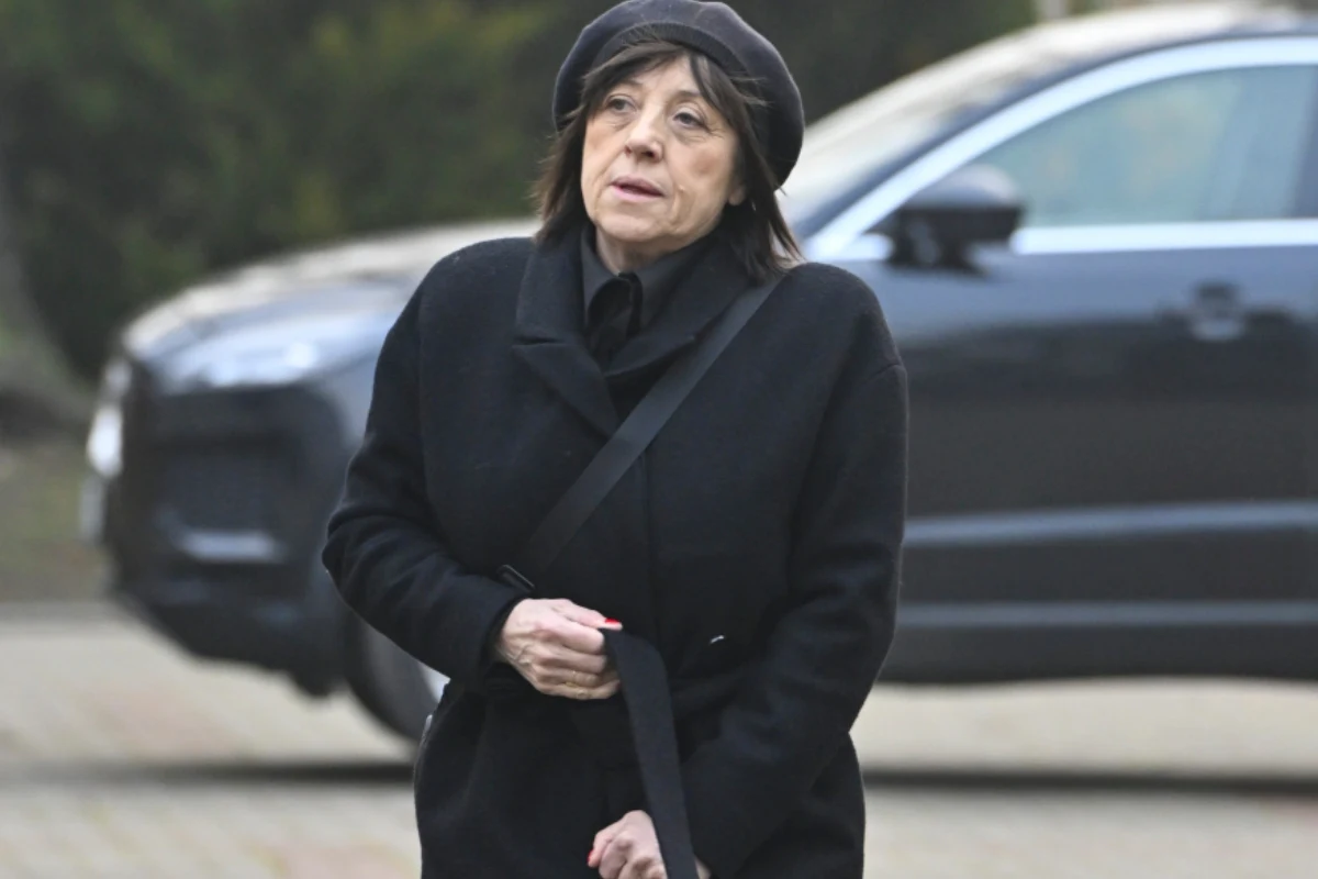 Starsza kobieta w czarnym płaszczu i berecie idzie ulicą, w tle widoczny samochód.