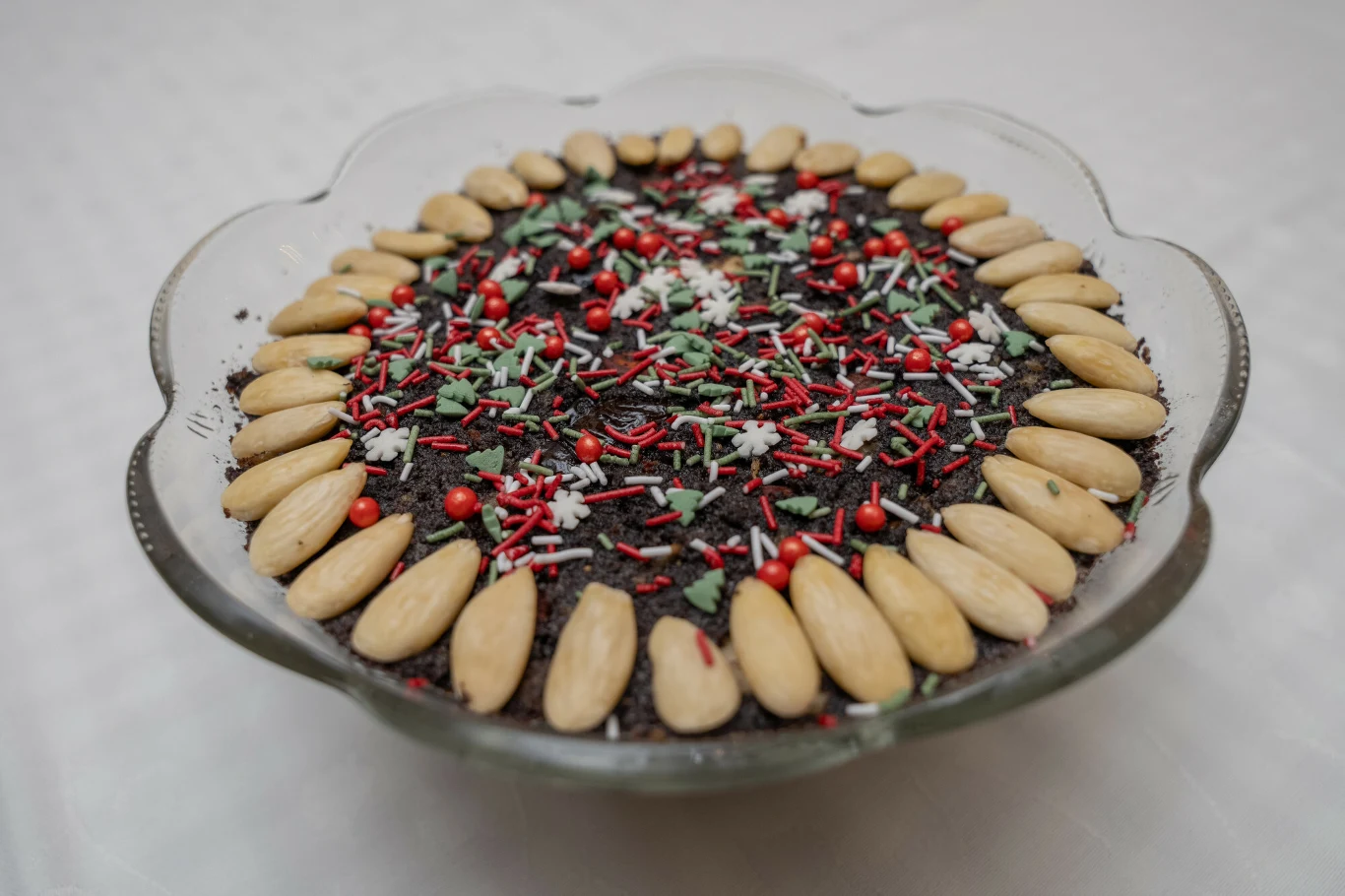 Dekorowany deser w szklanej misie, ozdobiony na wierzchu całymi migdałami ułożonymi w okrąg, kolorowymi posypkami oraz czerwonymi koralikami cukrowymi na ciemnej warstwie, całość na jasnym, jednolitym tle.