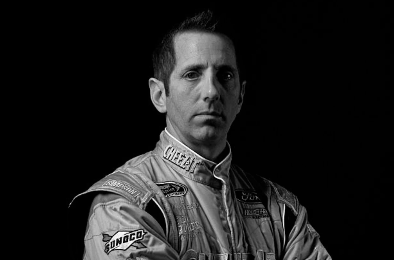 Nie żyje legenda NASCAR - Greg Biffle. Zginął w katastrofie odrzutowca w USA. Portret mężczyzny w kombinezonie kierowcy wyścigowego, stojącego z założonymi rękoma na ciemnym tle, wyraz twarzy poważny, liczne naszywki sponsorów na kombinezonie.
