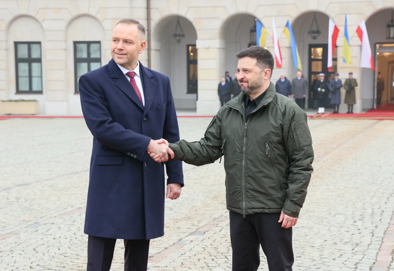 Dwóch mężczyzn ściska sobie dłonie na tle reprezentacyjnego budynku z arkadami, w tle widoczne są flagi narodowe oraz kilka osób w mundurach stojących przy wejściu.
