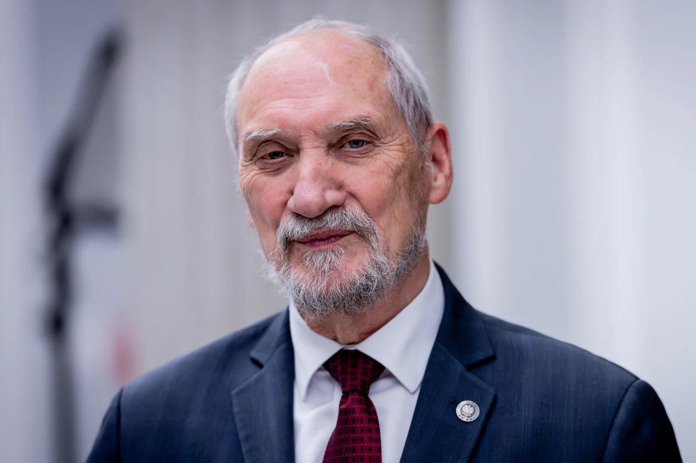 Sejmowa komisja podjęła decyzję w sprawie Antoniego Macierewicza. Odpowie za wykroczenia drogowe. Starszy mężczyzna z siwą brodą i krótkimi włosami, ubrany w granatowy garnitur, białą koszulę i ciemnoczerwony krawat, stoi na tle rozmytego jasnego wnętrza.