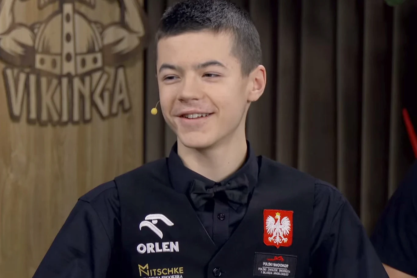 Młody mężczyzna w eleganckim ubraniu z kamizelką ozdobioną naszywkami sponsorskimi oraz godłem Polski, uśmiecha się podczas rozmowy w studiu telewizyjnym, w tle widoczne drewniane dekoracje i napis VIKINGA.