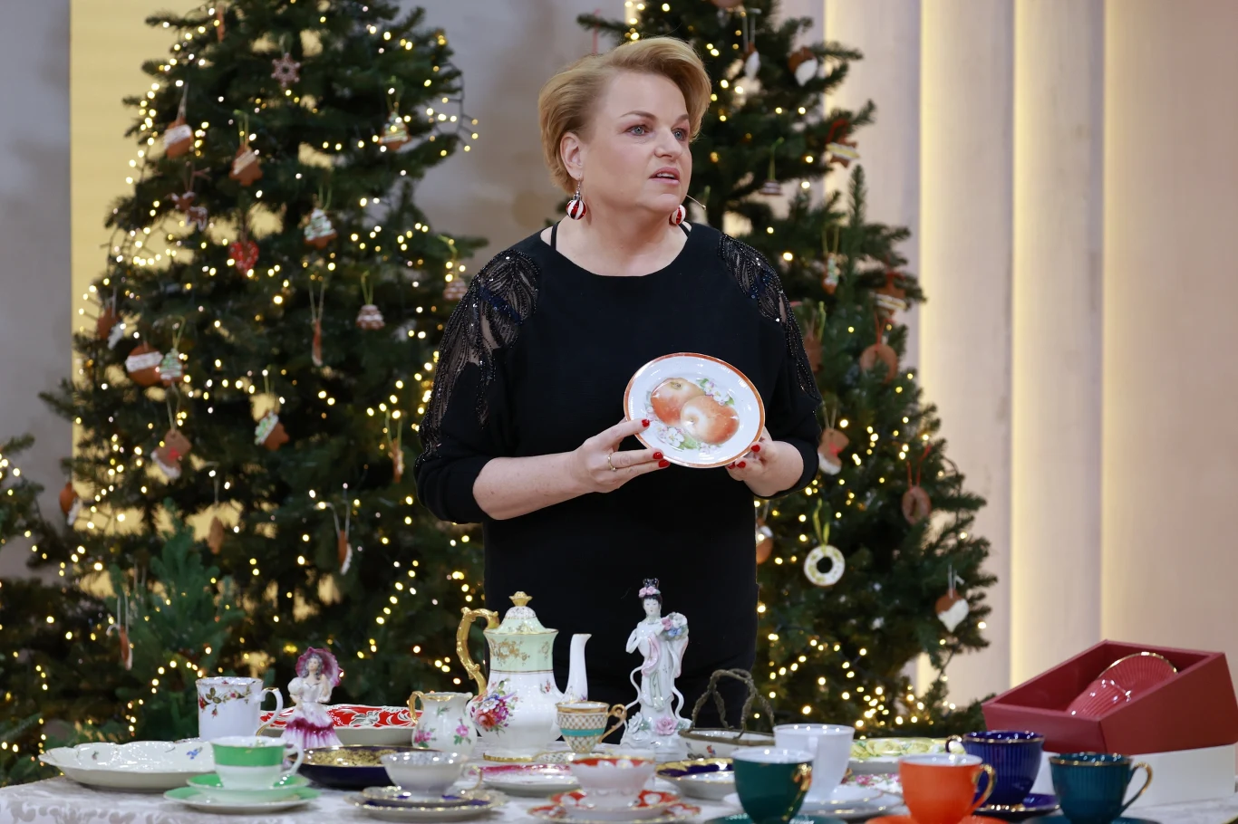 Katarzyna Bosacka zdradziła kilka trików, jak kupić dobre uszka i pierogi Kobieta w eleganckim stroju prezentuje porcelanowy talerz z wzorem, stojąc przy stole pełnym porcelany i filiżanek; w tle ozdobione choinki z lampkami świątecznymi.