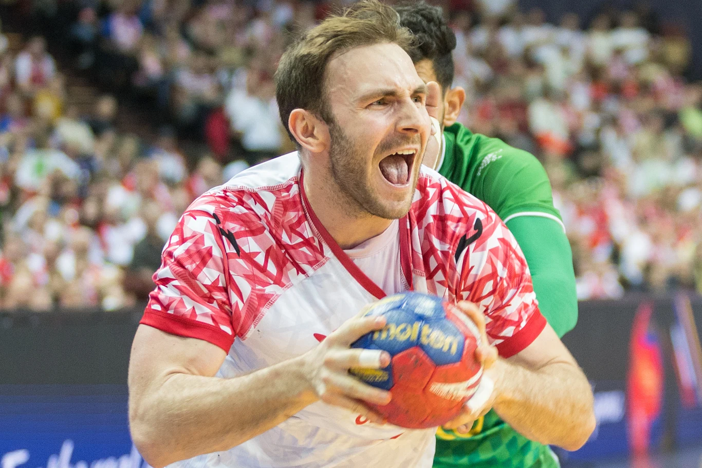 W piątek ogłoszono listę powołanych zawodników na ME. Co z Arkadiuszem Moryto? Zawodnik w biało-czerwonej koszulce z piłką do ręcznej w dłoniach, w trakcie dynamicznej akcji, wyrażający silne emocje. W tle widoczni inni zawodnicy i rozmyta publiczność.