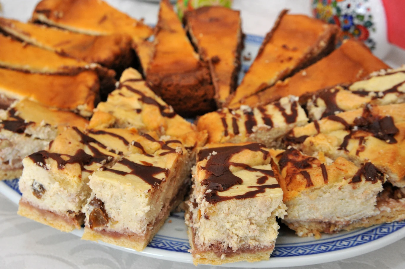 Domowy sernik był dla Tomasza największym rarytasem, gdy był małym chłopcem Kawałki domowego ciasta sernikowego z dodatkiem rodzynek i polewą czekoladową ułożone na ozdobnym talerzu, w tle fragmenty innych kawałków ciasta.