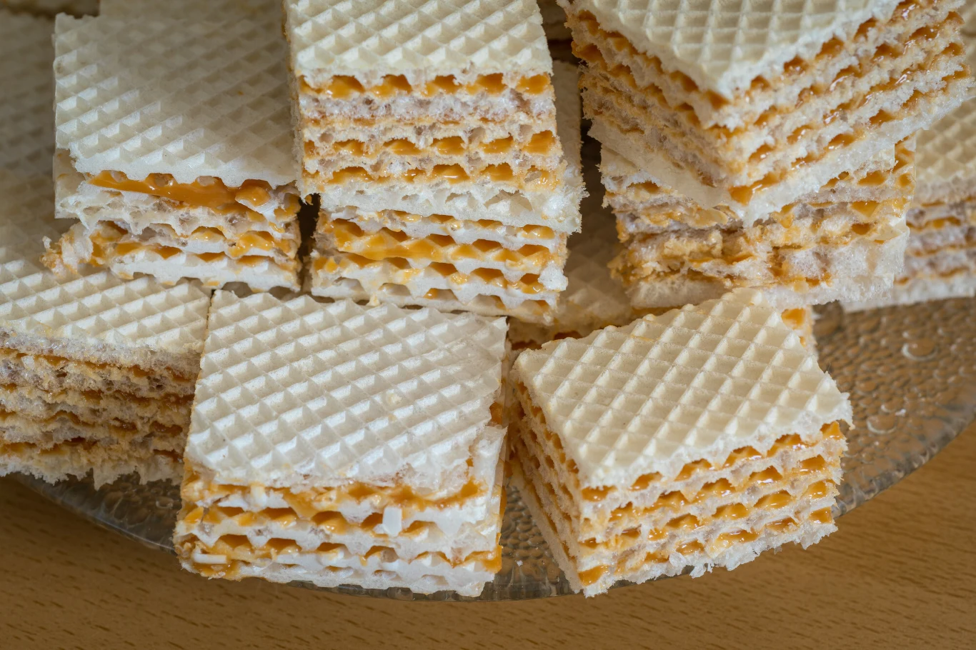 Wafle przekładane słodką masą, a potem krojone przez mamę są ulubionym wspomnieniem z dzieciństwa Ewy Kwadratowe wafle przełożone kremem o żółtym kolorze, ułożone na szklanym talerzu. Dominują regularne, warstwowe struktury i chrupiąca faktura wierzchniej warstwy.