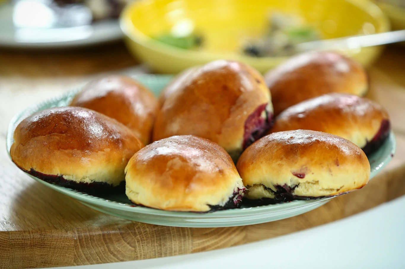 Zapach i smak jagodzianki od taty pamiętam do dziś Kilka apetycznych, złocistych bułeczek z owocowym nadzieniem ułożonych na jasnym talerzu, w tle rozmazane naczynia kuchenne.