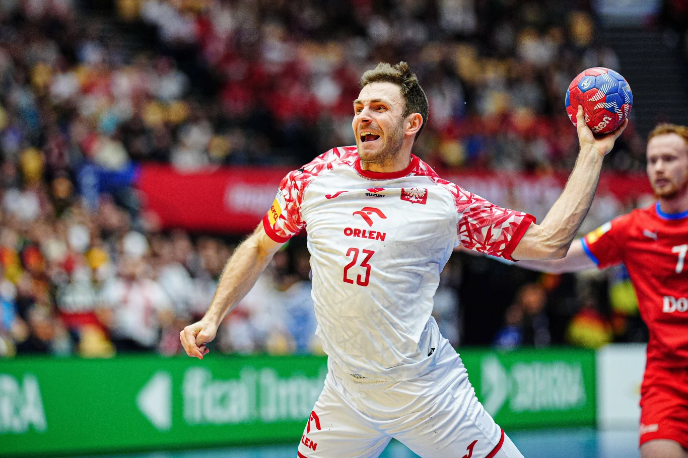 Arkadiusz Moryto Zawodnik drużyny piłki ręcznej w białym stroju z numerem 23 przygotowuje się do rzutu piłką, obok niego widoczny jest rywal w czerwonym stroju, w tle rozmyta publiczność na trybunach oraz bannery reklamowe.