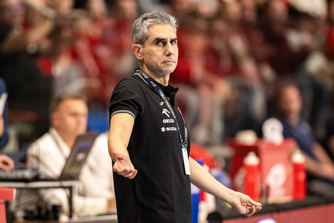 Jota Gonzalez Mężczyzna o siwych włosach w czarnej koszulce sportowej stoi na tle rozmytego tłumu na trybunach, patrząc poważnie przed siebie z szeroko rozłożonymi rękami, w tle widoczne są elementy hali sportowej oraz kibice.