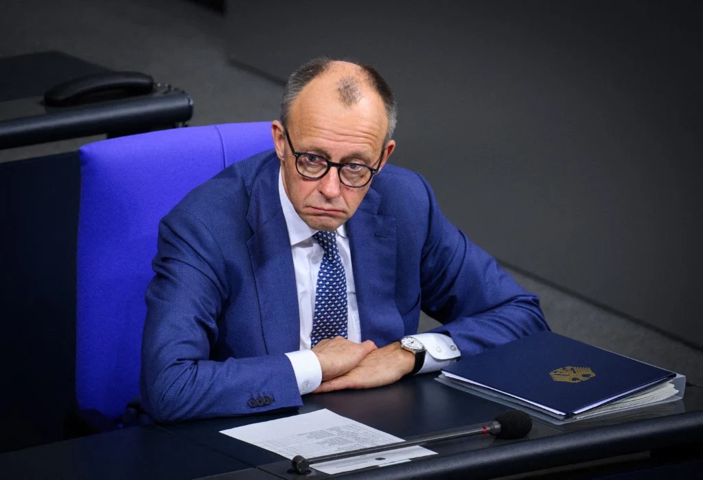 "Die Welt" o szczycie UE: Czarny dzień dla Merza Mężczyzna w garniturze siedzący przy stole z niebieskim segregatorem i dokumentami, z poważnym wyrazem twarzy, patrzy przed siebie w jasnym oświetleniu, na tle ciemnej sali.