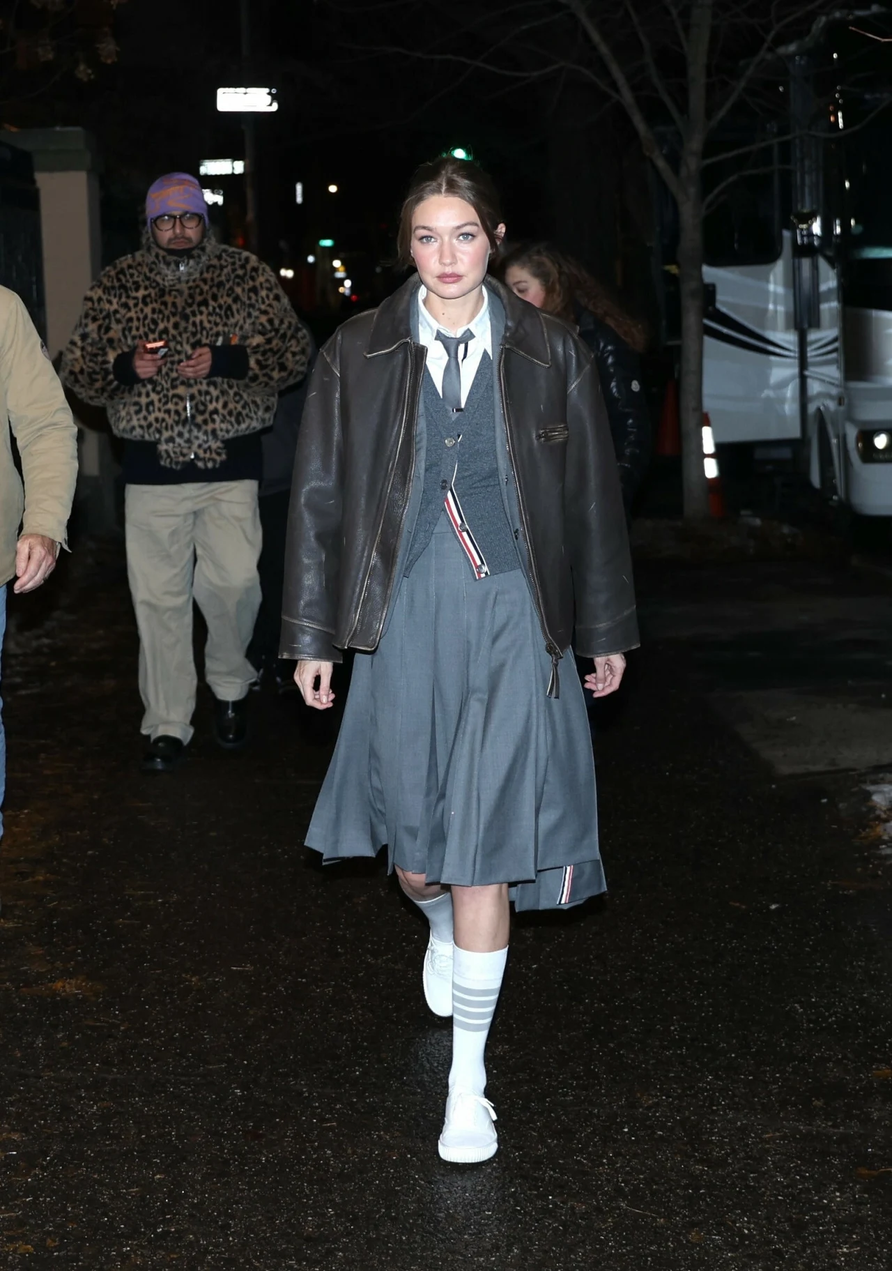 Gigi Hadid w oryginalnej stylizacji Młoda kobieta idzie pewnym krokiem ulicą w szarej sukience, swetrze, białych butach i skarpetkach do połowy łydki, ubrana w brązową skórzaną kurtkę, z tyłu widać kilka innych osób w różnych ubraniach.
