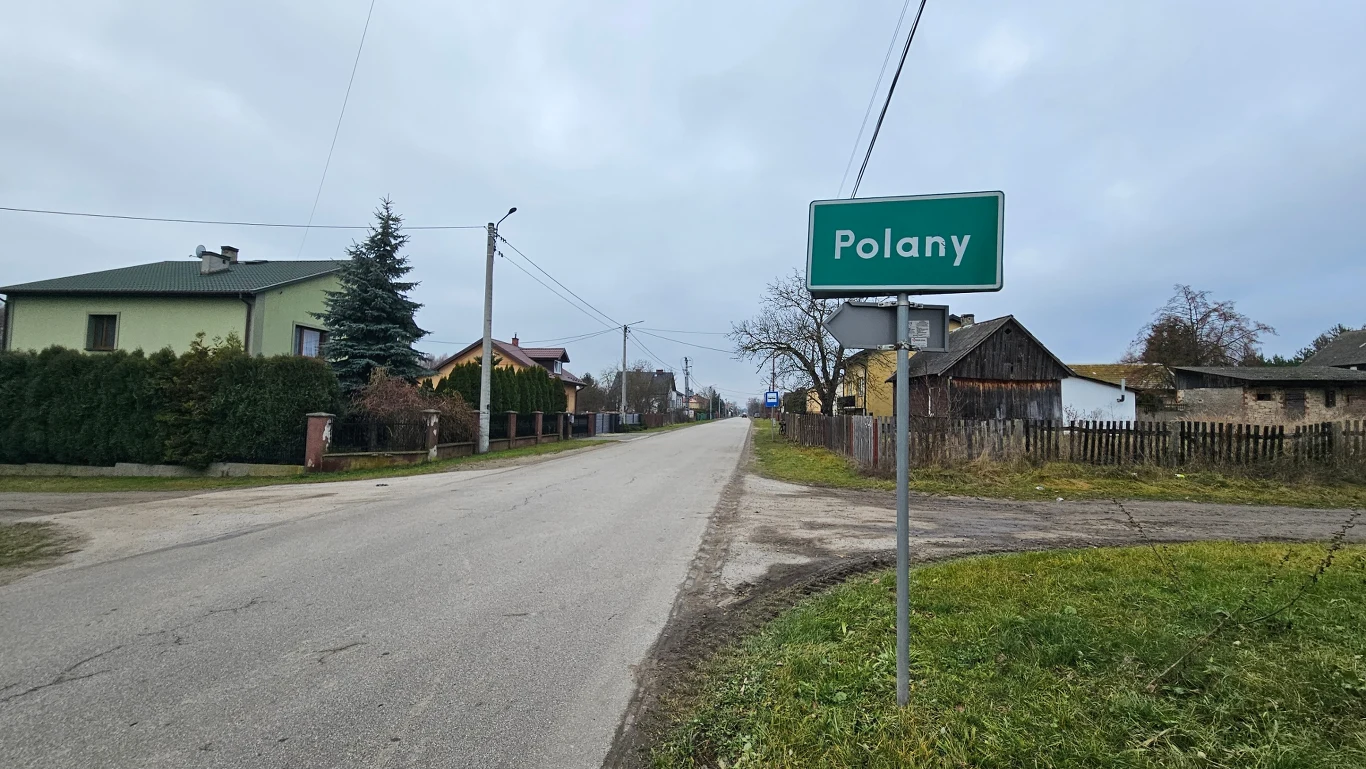 Wiejska droga prowadząca przez miejscowość Polany, po obu stronach drogi domy mieszkalne, ogrodzenia oraz zieleń. Po prawej stronie widoczna tablica z nazwą miejscowości. Niebo zachmurzone, wokół spokój i brak ruchu ulicznego.