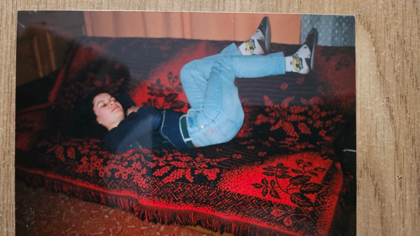 Kobieta leżąca na plecach na czerwono-czarnej kanapie z ozdobnym wzorem, ma ugięte kolana i nosi jasne dżinsy oraz skarpetki z kolorowym motywem.