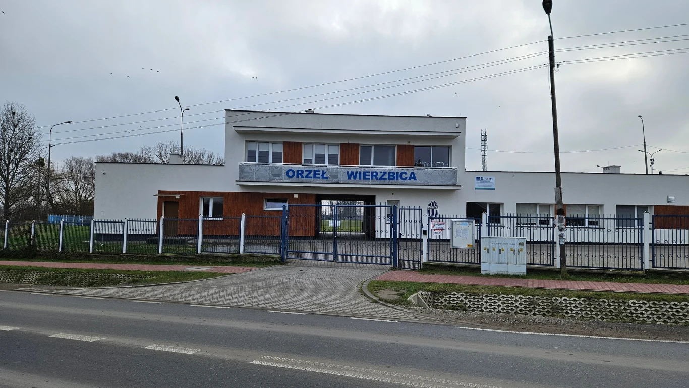 Nowoczesny budynek klubu sportowego z szyldem Orzeł Wierzbica, otoczony ogrodzeniem, z zamkniętą bramą oraz widocznym trawnikiem i elementami chodnika przed wejściem.