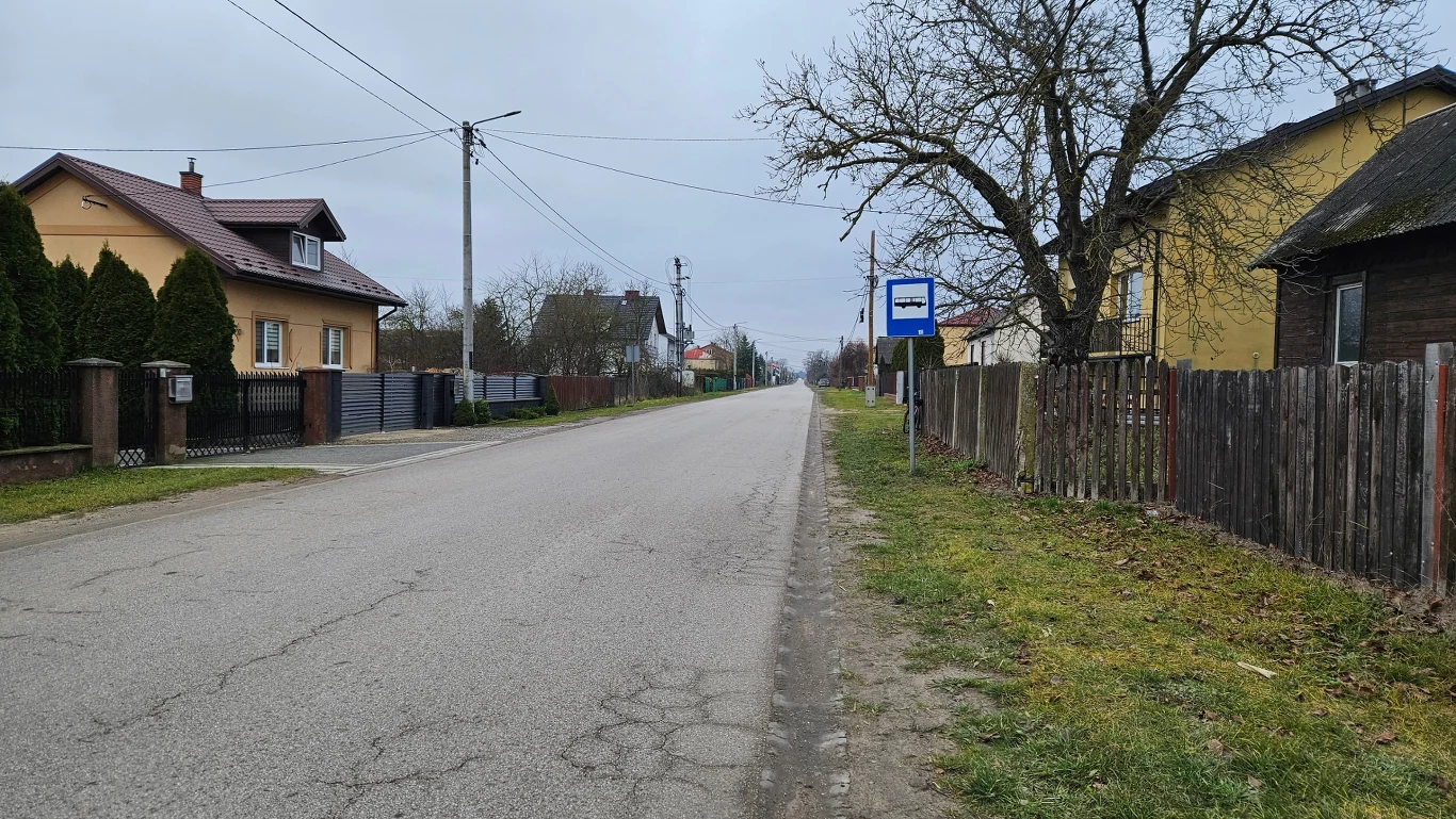 Pusta asfaltowa droga biegnąca przez wiejską miejscowość, po jednej stronie otoczona drewnianymi i metalowymi ogrodzeniami, po drugiej stronie widoczne domy jednorodzinne, przystanek autobusowy oraz przydrożne drzewa.