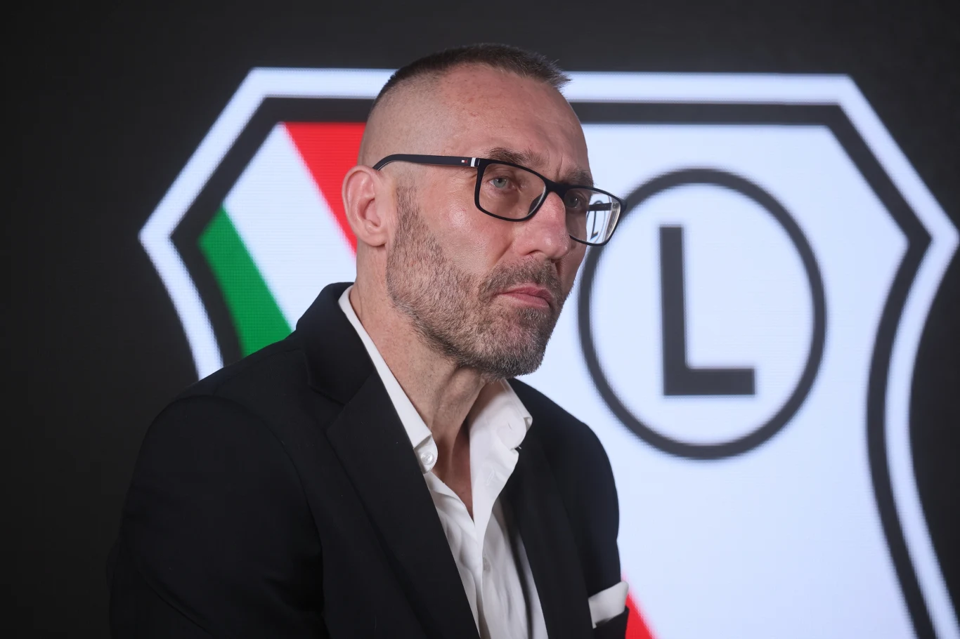 Mężczyzna w garniturze i okularach siedzący na tle dużego, graficznego logotypu w nowoczesnych barwach, z wyraźną literą L po prawej stronie.