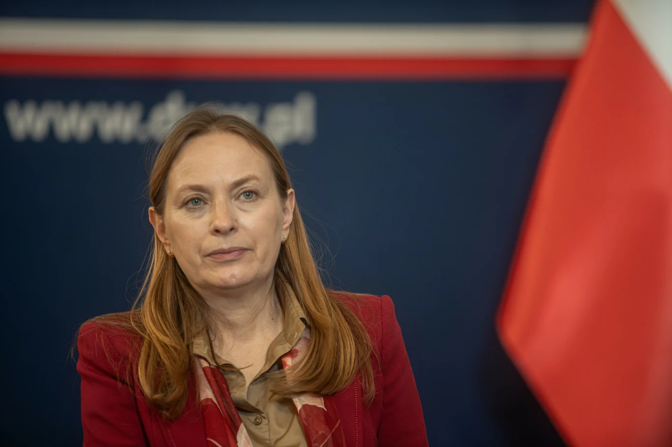 Nowa inicjatywa wspiera wynajem mieszkań w Polsce, ma łączyć ludzi z pustostanami (zdj. ilustracyjne) Kobieta w średnim wieku z długimi brązowymi włosami, ubrana w bordową marynarkę i beżową bluzkę, stoi na tle ciemnoniebieskiej ściany z fragmentem polskiej flagi oraz częściowym napisem internetowym.